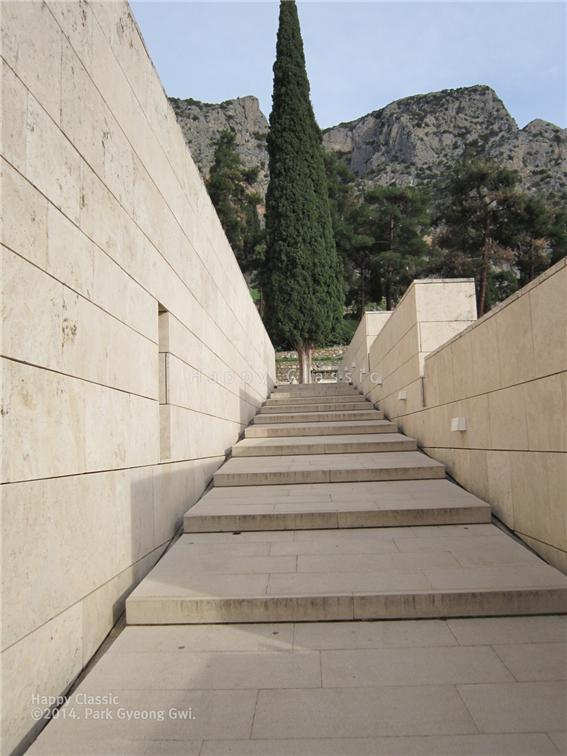 델피 고고학 박물관 1층 입구로 오르는 계단이다. 장애인들은 0층(한국의 1층에 해당)으로 바로 입장할 수 있다. 파르나소스의 산정의 절벽과 사이프러스 나무가 조화를 이룬다. 마치 ‘신성한 길’을 오르듯 한 계단 한 계단을 오르며 가슴이 설렌다. ⓒ박경귀